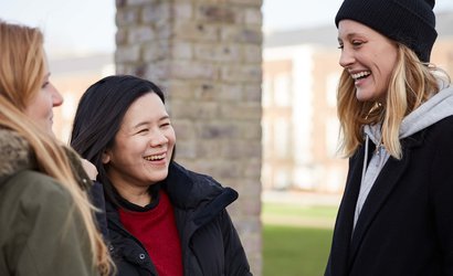 Three women stand in conversation in front of a brick pillar with a blurred building in the background.