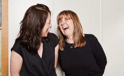 Two people standing side-by-side looking happy and laughing together against a wall in a house.