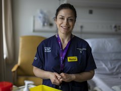 A close up of a healthcare professional wearing blue scrubs, smiling at the camera with hospital equipment out of focus in the background.