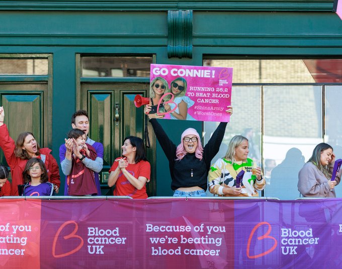 A group of people stand behind partitions at the side of the London Marathon. They are clapping and cheering for the runners taking part in the event. They have clapper boards and a woman with pink hair is holding up a sign that says 'Go Connie!'. It is a sunny day and some of them are wearing Blood Cancer UK t-shirts.
