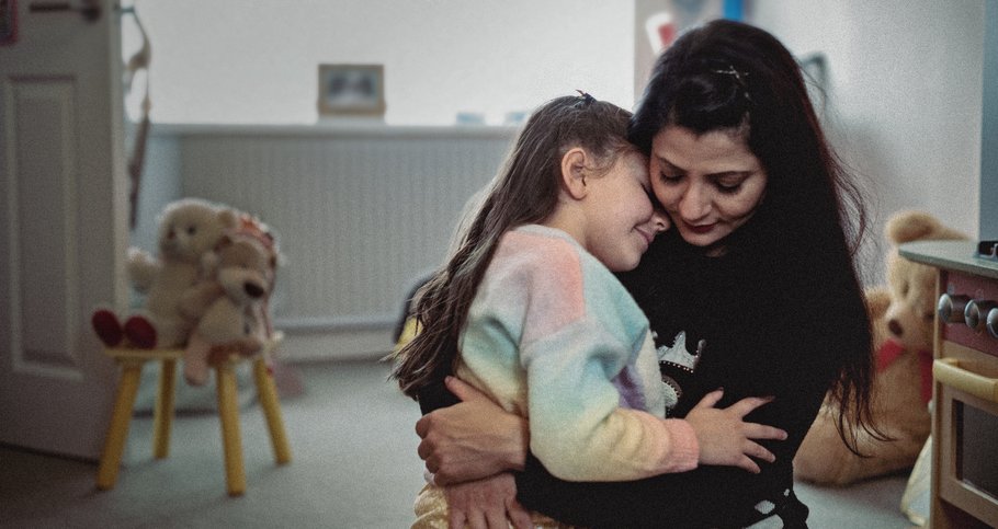 A young girl sits on her mother's lap and gives her a hug. They are sitting in a family home and toys are visible in the background.