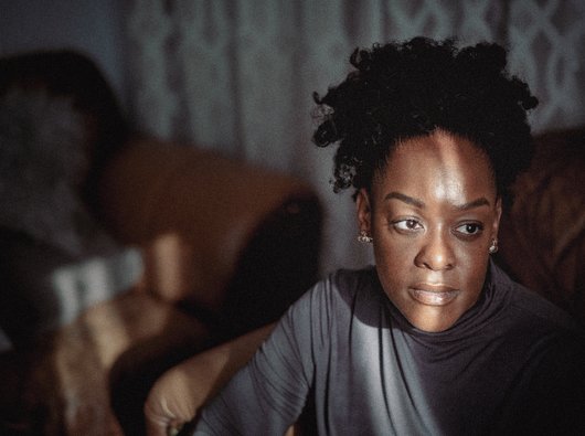 A black woman sits in her family home. She is looking into the distance on her left. There is a ray of light across her face.