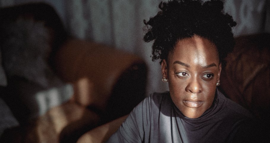 A black woman sits in her family home. She is looking into the distance on her left. There is a ray of light across her face.