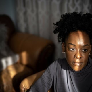 A close up of a black girl standing near a window, the curtains causing light and shadows to create a pattern across her face.