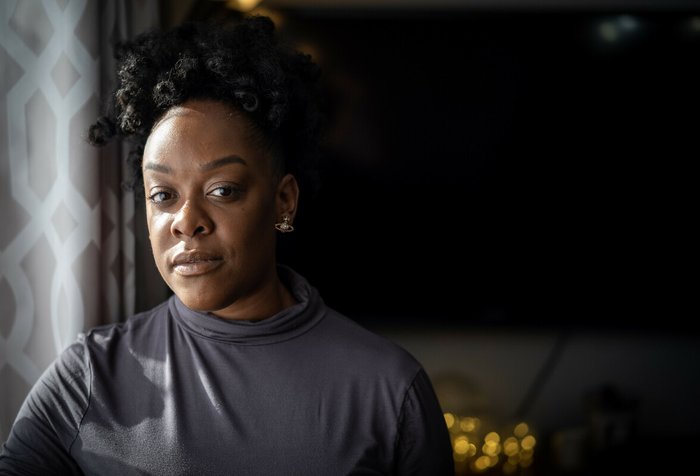 A close up of a black girl looking straight into the camera, light shining on her face with the background surrounded in shadows.