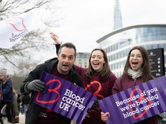 Blood Cancer UK staff members cheering and smiling at an event, holding flags and banners.