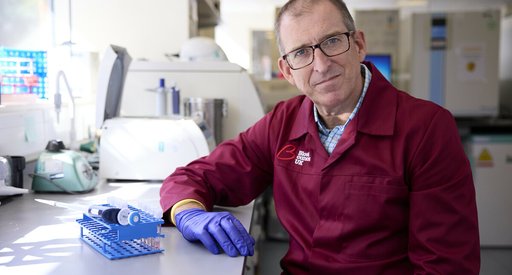 A man wearing glasses and a red Blood Cancer UK lab coat sat in a science lab