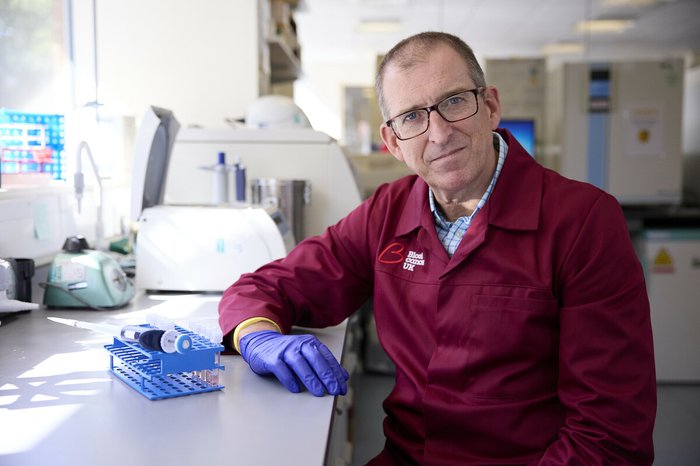 A man wearing glasses and a red Blood Cancer UK lab coat sat in a science lab