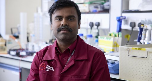 A close up of a researcher wearing a Blood Cancer UK, maroon lab coat, looking straight into the camera with a serious expression.