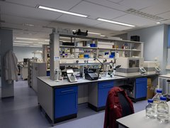 Inside a research lab, work bench with lab equipment and chair with a red Blood Cancer UK lab coat hung over the back.
