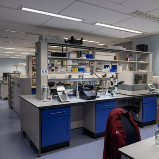 Inside a research lab, work bench with lab equipment and chair with a red Blood Cancer UK lab coat hung over the back.