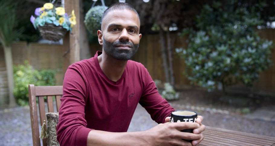 A man is sitting at a garden table, looking at the camera and holding a cup of tea in both hands. He is smiling slightly, and seems thoughtful. There are fairy lights hanging above him and a basket of flowers in the background.
