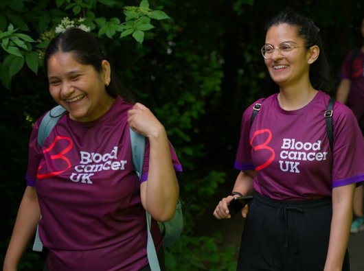 Two smiling women on a charity walk through woodland. They have backpacks on and branded Blood Cancer UK tshirts. Just out of focus behind them, two more walkers can be seen.