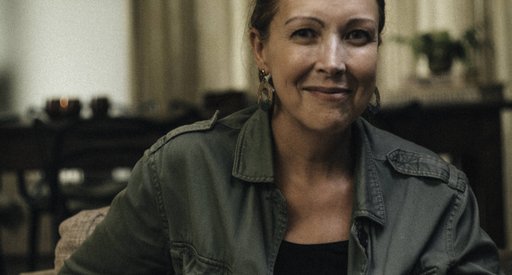 A middle aged woman looks straight at the camera with a slight smile; she is surrounded by a darkly lit room.