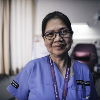 A close up of a healthcare professional wearing blue scrubs and smiling slightly at the camera in a dimly lit room.