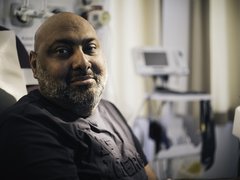 A close up of a man in a hospital bed, looking straight into the camera with a slightly apprehensive smile.
