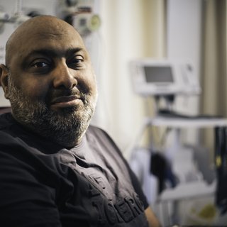 A close up of a man in a hospital bed, looking straight into the camera with a slightly apprehensive smile.