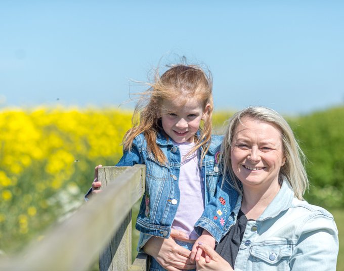 Kirsty holding her daughter up by a fence against a yellow field in the sunshine. They are both smiling with blue sky behind.