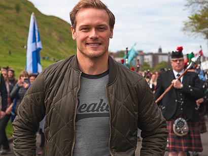 Sam Heughan, Scotland President of Blood Cancer UK, standing in front of the camera with a pipe band in the background.