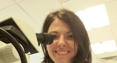 A lady in a science lab, wearing a white lab coat, smiling next to a microscope.