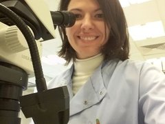 A lady in a science lab, wearing a white lab coat, smiling next to a microscope.