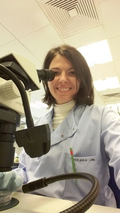 A lady in a science lab, wearing a white lab coat, smiling next to a microscope.