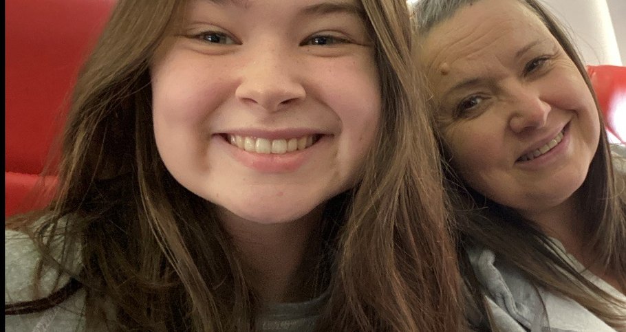 Sophie smiles at the camera. Her head and shoulders are in shot and it looks like she is sat on a train. Her mum is next to her, also smiling at the camera.