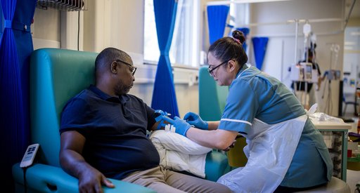 A male patient and a female nurse on day unit. She is taking a blood test from his left arm. They are both sitting down.