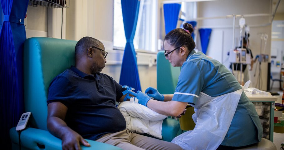 A male patient and a female nurse on day unit. She is taking a blood test from his left arm. They are both sitting down.