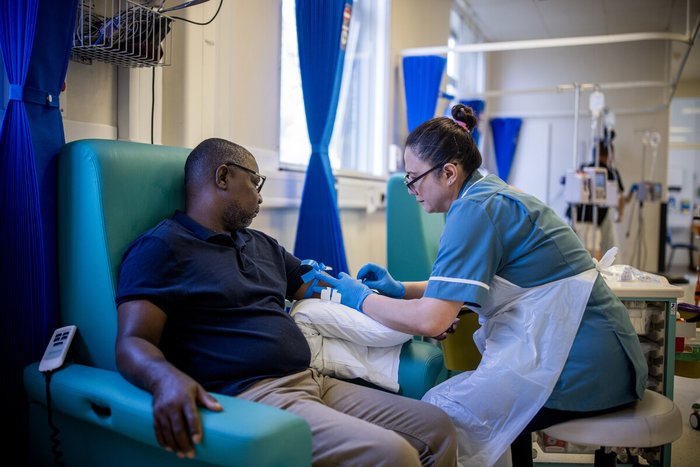 A male patient and a female nurse on day unit. She is taking a blood test from his left arm. They are both sitting down.