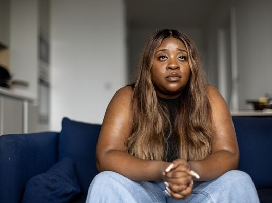 A woman sat on a sofa with her hands clasped. She's looking thoughtful and sad.