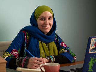 A woman in a headscarf sits at a laptop. She is holding a pen and smiling at the camera.
