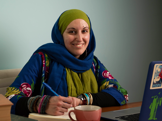 A woman in a headscarf sits at a laptop. She is holding a pen and smiling at the camera.