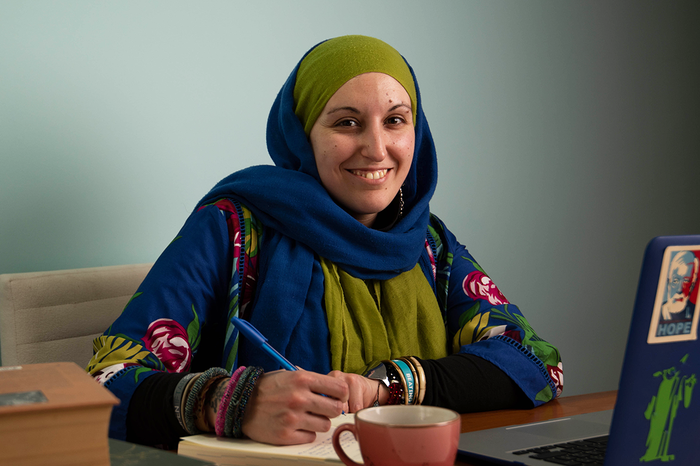 A woman in a headscarf sits at a laptop. She is holding a pen and smiling at the camera.