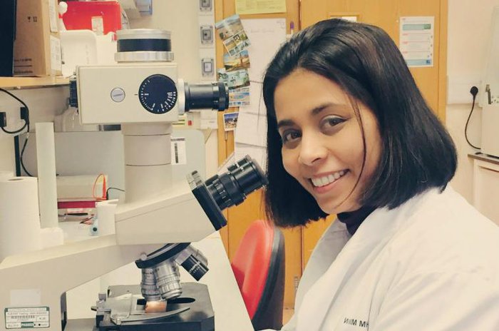 Anndita Roy - a researcher - in a lab looking directly at the camera with a microscope