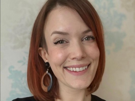 Portrait photo of a member of staff with red hair and wearing a black top. They are smiling at the camera.