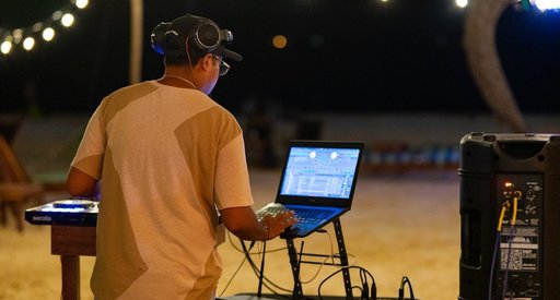 A DJ at an event. He has his back to us and is looking at a lap top. There's a speaker and festoon lights in the background. He has dark hair and brown skin and is wearing a cap with glasses and headphones on his head.