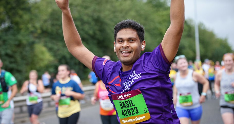 A male runner holds his arms up in the air making peace signs and smiling at the camera. He's wearing a Blood Cancer UK branded t-shirt and wearing in-ear headphones.