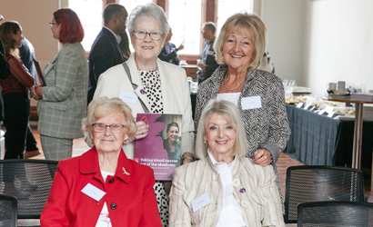 A group of four women at an event sit facing the camera and smiling. A lady in glasses at the back of the group is holding a copy of the Blood Cancer Action Plan and smiling.