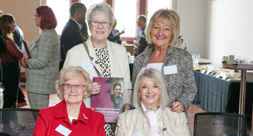 A group of four women at an event sit facing the camera and smiling. A lady in glasses at the back of the group is holding a copy of the Blood Cancer Action Plan and smiling.