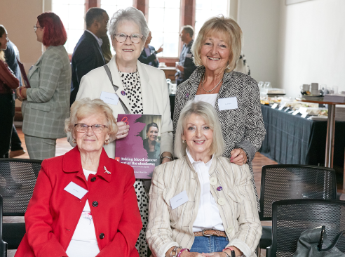 A group of four women at an event sit facing the camera and smiling. A lady in glasses at the back of the group is holding a copy of the Blood Cancer Action Plan and smiling.