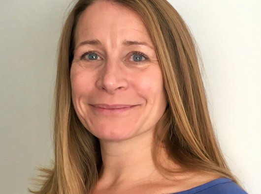 Head-and-shoulders portrait of a person with long light brown hair, wearing a blue top and looking at the camera with a slight smile, against a plain light-coloured background.
