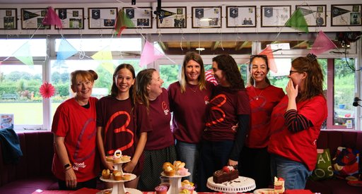 A group of smiling and laughing people stand around a table of cakes in a community hall.