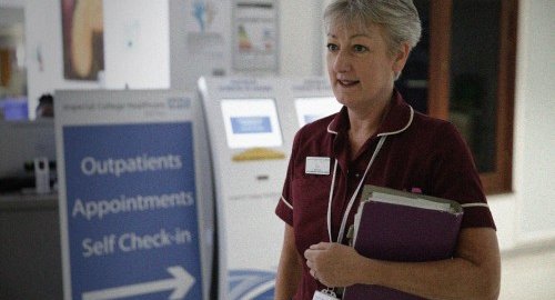 A nurse stands at an out patient clinic. She has short hair and is holding a clipboard.
