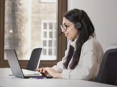 A woman with dark hair and glasses works on a support line. She is looking at a laptop and has a headset on.