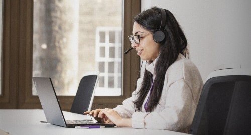 A woman with dark hair and glasses works on a support line. She is looking at a laptop and has a headset on.