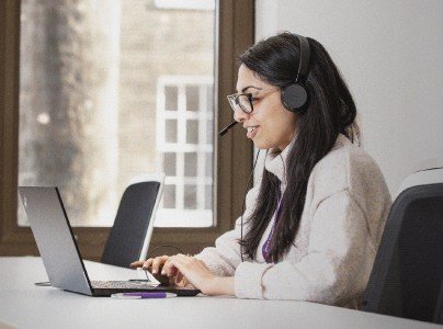 A woman with dark hair and glasses works on a support line. She is looking at a laptop and has a headset on.