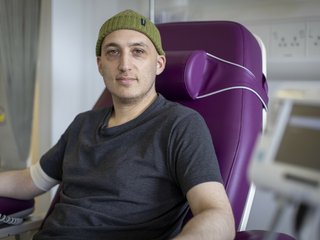 A man having treatment in hospital. He is sitting in a purple hospital bed, wearing a grey t shirt and a woollen beanie hat.