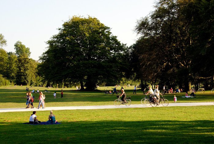 A wide shot of a park with people walking, sitting and cycling in it.