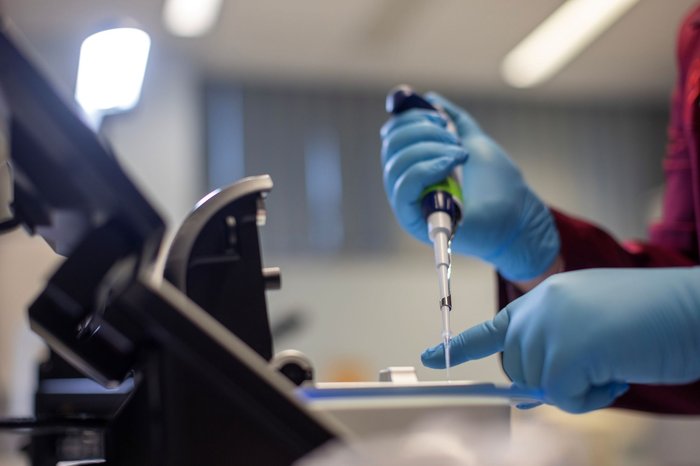 Laboratory image in which a pair of gloved hands use a syringe to inject liquid onto a microscope.